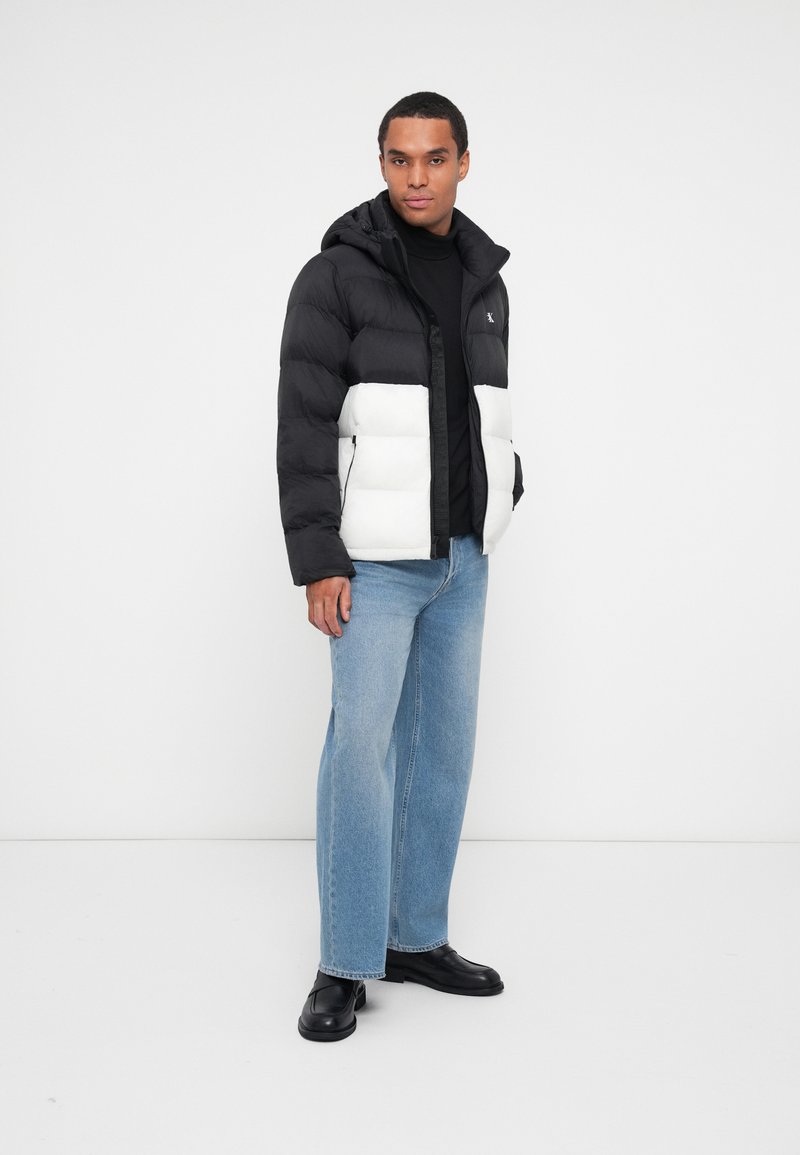 Schwarz-weiße Steppjacke mit Kapuze, Reißverschluss und zwei seitlichen Taschen. Das Model trägt hellblaue Jeans und schwarze Schuhe.