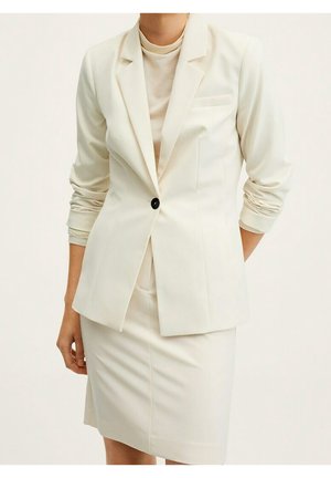 Vrouw die een crème blazer draagt met één zwarte knoop en een bijpassende kokerrok tot op de knie, mouwen licht gerimpeld.