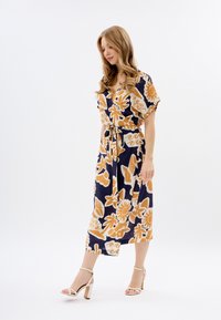 Robe bleu marine à motifs floraux avec des manches courtes et une taille ceinturée. Présente une jupe fluide de longueur midi, portée avec des sandales à talons blocs blanches.