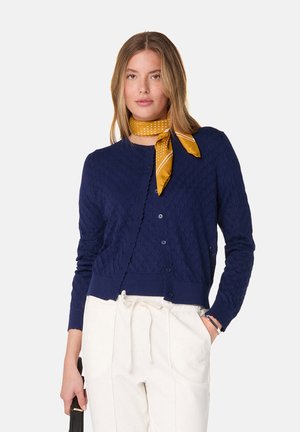 Cardigan en tricot bleu marine avec motif en losanges, poignets et ourlet côtelés, encolure ronde et boutons assortis. Porté par-dessus un pantalon crème.