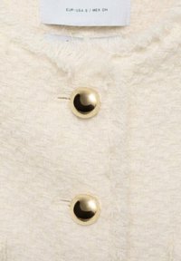 Cream textured fabric with a boucle pattern, featuring two gold-tone round buttons and a clothing label showing size and origin.
