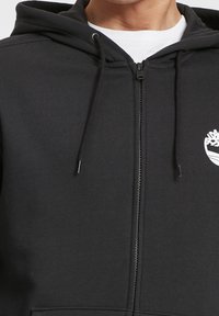 Sudadera con cremallera negra hecha de tejido suave, con una cremallera frontal, dos cordones y un logo blanco en el lado izquierdo del pecho.