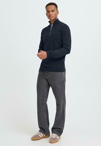 Sötét tengerészkék félcipzáros pulóver hosszú ujjakkal, texturált anyagból, szürke farmernadrággal és fehér csíkos bézs sportcipővel párosítva.