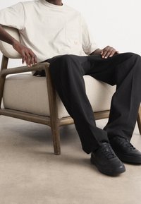 Beige bomullströja med bröstficka, kombinerad med svarta vida byxor och svarta sneakers, sittande på en ljusklädd fåtölj.