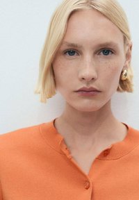 Orange cardigan med rund halsringning och knappstängning. Slät textur och korta ärmar. Modellen har kort blont hår och bär guldörhängen.