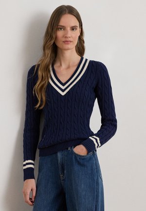 Mujer con cabello largo y ondulado, vistiendo un suéter azul marino de punto trenzado con rayas blancas en el cuello en V y los puños, combinado con jeans de mezclilla azul, con la mano en el bolsillo.