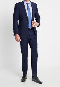 Námořnický modrý oblek se přiléhavým sakem a rovnými kalhotami, doplněný o světlou pruhovanou košili a modrou kravatu. Černé kožené boty.