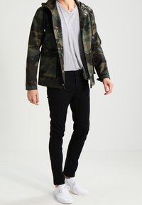 Chaqueta de camuflaje en tonos oscuros, con capucha, botones y bolsillos frontales, llevada sobre una camiseta gris y pantalones slim-fit negros.