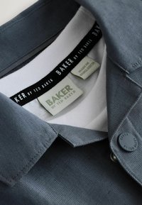 Gros plan sur une chemise grise à col avec étiquettes de marque Ted Baker et bouton, montrant la texture du tissu et une étiquette "Fabriqué au Bangladesh".