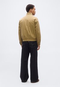 Άνδρας με μπεζ μπουφάν και σκούρο παντελόνι φαρδιάς γραμμής στέκεται με την πλάτη προς τα εμπρός, μπροστά σε λευκό φόντο.