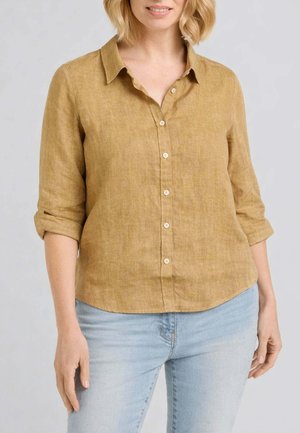 Donna che indossa una camicia gialla senape con bottoni e maniche arrotolate, e jeans azzurri, in piedi davanti a uno sfondo neutro.