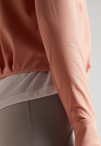 Top atlético ligero de color durazno con tela perforada, mangas largas y acentos reflectantes en el brazo izquierdo. Superpuesto sobre una parte inferior de malla gris.