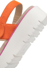 Oranges Wildledersandalette mit einem breiten Riemen und weißer Sohle. Die Sohle hat einen strukturierten Grip und einen rosa Akzent entlang des Randes.