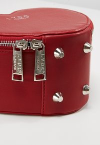 Rode nepleer ronde tas met zilverkleurige hardware, voorzien van dubbele ritsen en metalen studs langs de bodem voor extra detail.