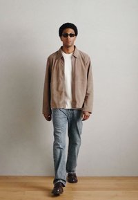 Világosbarna velúr dzseki, fehér póló, világoskék farmer és barna cipők. Kerek napszemüveg teszi teljessé a laza öltözéket. Fa padló háttere.