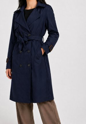 Kvinde iført navy dobbeltbrystet trenchcoat med bælte og detaljer i tan spænde, kombineret med brune vidde bukser mod en ensfarvet baggrund.