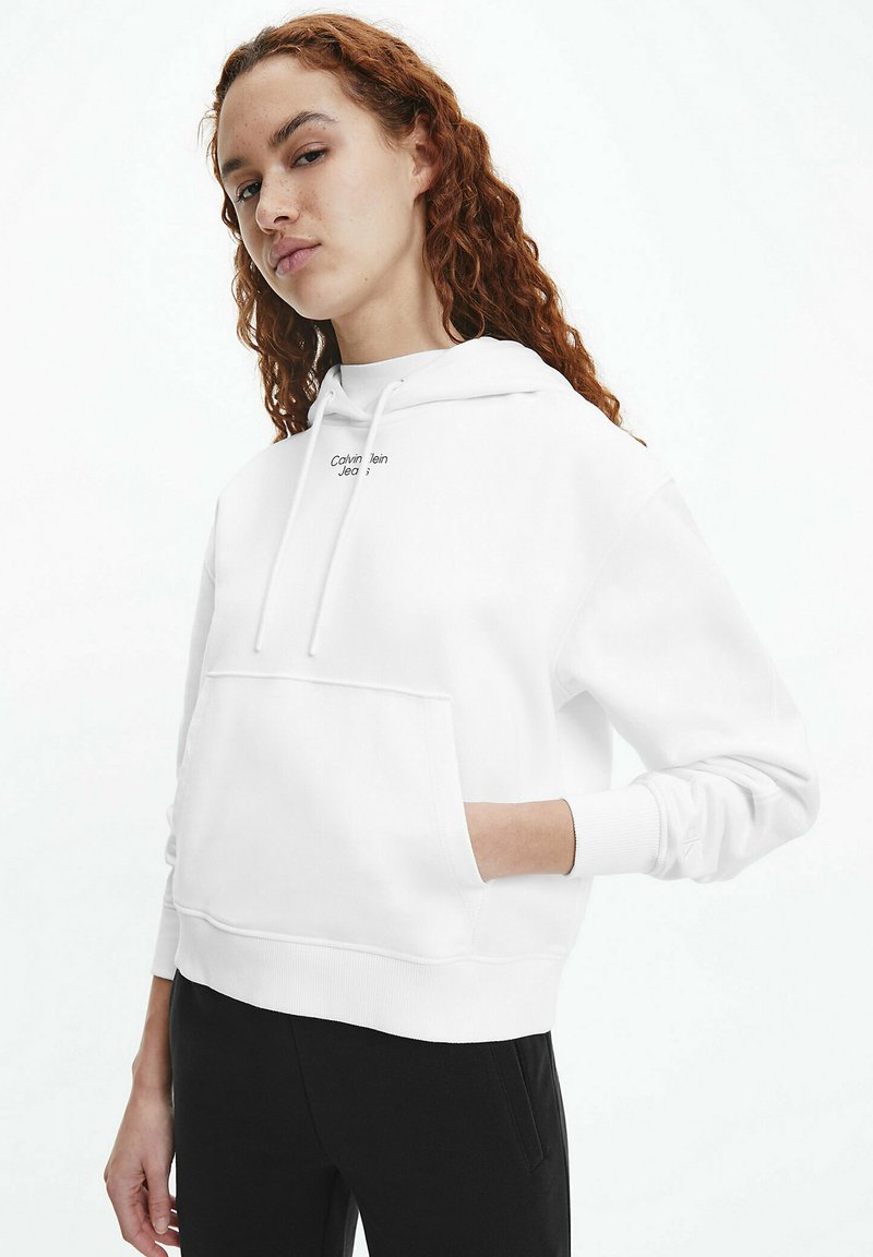 Sudadera blanca con bolsillo frontal, capucha con cordón y logo de "Calvin Klein Jeans" en negro. Presenta puños y dobladillo acanalados.
