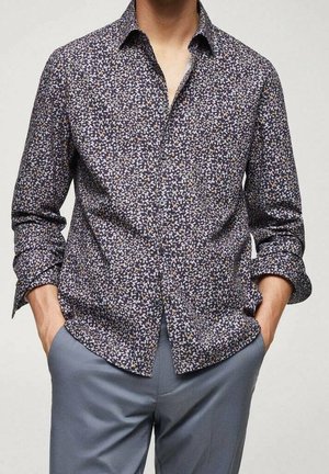 Homme portant une chemise à manches longues sombre à motifs avec les manches retroussées et un pantalon gris clair, les mains dans les poches.