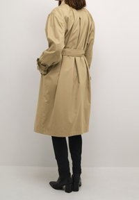 Beżowy płaszcz trenczowy z plisą z tyłu i paskiem, z długimi rękawami i średniej długości krojem. W zestawie z czarnymi dopasowanymi spodniami i botkami na obcasie.