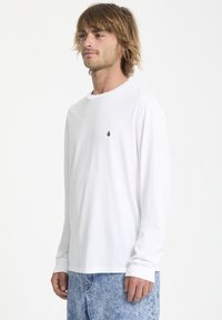 Langärmliges weißes T-Shirt aus Baumwolle mit rundem Hals und gerippten Bündchen, das ein kleines schwarzes Logo auf der Brust trägt.