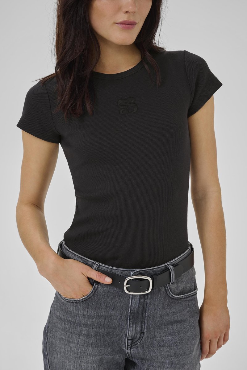 Naine kannab vormis musta lühikeste varrukatega tärklisega toppi, millel on peen logo, sisenõgitud tuhmunud hallidesse teksapükstesse koos musta nahkvööga.