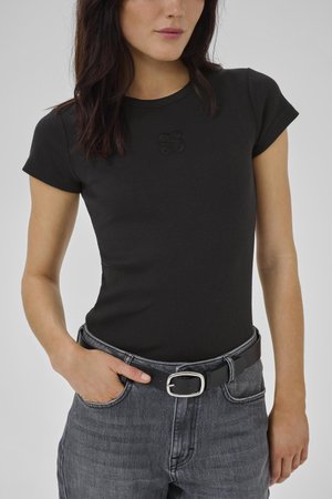 Vrouw draagt een nauwsluitend zwart shirt met korte mouwen en een subtiel logo, ingestopt in vervaagde grijze spijkerbroek met een zwarte leren riem.