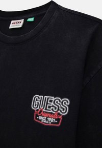 T-shirt in cotone nero con colletto rotondo. Presenta il logo ricamato "GUESS Originals" in bianco e rosso, con il dettaglio "Dal 1981" sottostante.