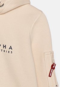 Felpa beige realizzata in tessuto morbido con stampa del logo, cappuccio con cordino e tasca con zip sulla manica con un tirante rosso.