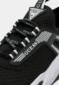 Μαύρο αθλητικό παπούτσι με υφασμένη επάνω επιφάνεια, λευκές λεπτομέρειες και κορδόνια με το λογότυπο της μάρκας. Διαθέτει λαστιχένια σόλα και τριγωνικό λογότυπο στη γλώσσα του παπουτσιού.