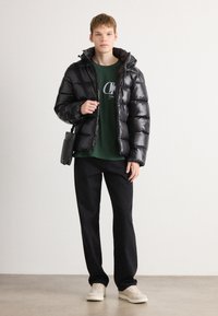 Zwarte pufferjack met een glanzende afwerking, capuchon en ritssluiting, gecombineerd met een groene logo-t-shirt, zwarte broek en lichtgekleurde sneakers.