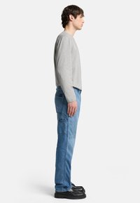 Maglietta grigia a maniche lunghe, pantaloni cargo in denim blu con tasche laterali e scarpe nere chunky, mostrati in un profilo laterale su uno sfondo bianco.
