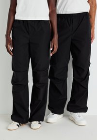 Δύο άτομα στέκονται δίπλα δίπλα φορώντας μαύρο φαρδύ παντελόνι, λευκά μπλουζάκια και λευκά αθλητικά παπούτσια, πάνω σε ένα απλό φωτεινό φόντο.