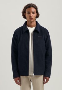 Navy zip-up jack in zachte, gestructureerde stof met een kraag en voorzakken. Kenmerkt een subtiele logo op de borst en manchetten.