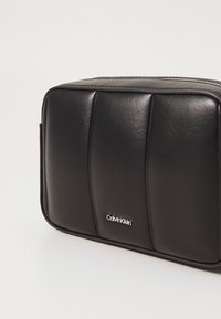Černá kožená rectangularní taška s texturovaným předním dílem, vertikálními panely a stříbrným logem na spodní části předu. Zapínání na zip.