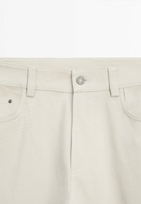 Pantalon beige clair avec ceinture avant, bouton métallique, passants de ceinture et détails de couture visibles.