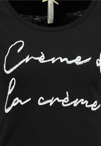 Černé bavlněné tričko s bílým flitrovým nápisem "Crème de la crème" v psacím písmu, kulatý výstřih. Měkká textura, neformální design.