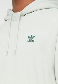 Közelkép egy személyről, aki világosszürke Adidas kapucnis pulóvert visel zöld háromlevéllogóval és a kapucnis zsinórokkal.