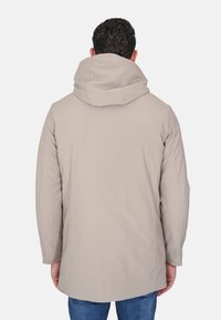 Uomo con giacca beige con cappuccio e jeans blu, visto di spalle contro uno sfondo semplice grigio chiaro.