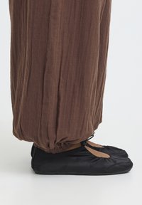 Ballerines noires avec des lanières élastiques portées avec un pantalon marron ample resserré aux chevilles sur fond blanc.