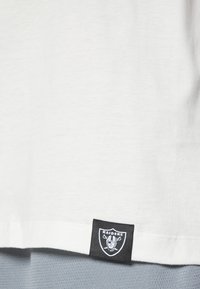 Camiseta blanca de algodón con un parche negro del logo "Raiders" en el dobladillo inferior. Textura suave, diseño casual y corte holgado.