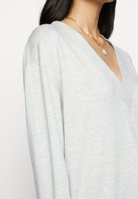 Suéter de manga larga en V de color gris claro, hecho de un material suave, con un corte relajado y una textura sutil. Un collar de cadena dorada añade un acento.