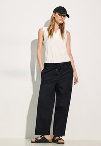 Top bianco senza maniche, pantaloni neri a gamba larga con vita elasticizzata e coulisse, cappello nero e sandali neri con base in sughero e accenti metallici.