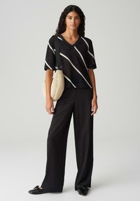 Maglione nero a V con una texture, decorato con strisce diagonali bianche, abbinato a pantaloni neri larghi e a una borsa a spalla intrecciata beige. Scarpe nere basse.