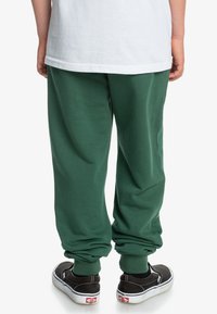 Pantalones de chándal verdes con puños elásticos, hechos de tejido suave, combinados con zapatos negros de tipo slip-on. El conjunto presenta un ajuste relajado y un diseño casual.