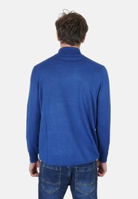 Maglione blu a maniche lunghe con collo alto, texture liscia e vestibilità ampia, abbinato a jeans in denim con dettagli di cuciture evidenti.
