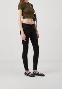 Top corto verde oliva, jeans negros ajustados y zapatos negros con tachuelas. Los accesorios incluyen un bolso negro y un collar de cadena plateado.