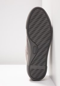 Semelle d'une chaussure décontractée grise avec une semelle extérieure en caoutchouc texturée, placée debout contre un mur d'angle blanc sur une surface blanche.