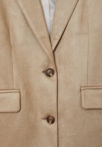 Blazer di tessuto beige con una trama liscia, dotato di due bottoni, rever a punta e due tasche frontali. Finitura leggermente lucida.