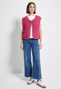 Fuchsia fleeceväst med knappfront, bärs över en vit t-shirt, matchad med vida blåjeans och vita sneakers med bruna accenter.