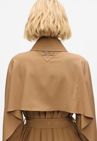 Indumento di colore marrone in stile mantella con colletto, caratterizzato da un dettaglio logo argentato e un elemento di gonna plissettata. Tessuto liscio con una leggera textura.